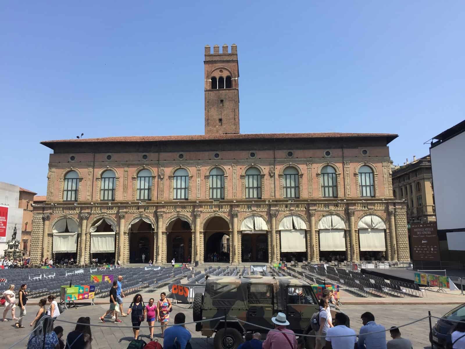Palazzo del Podestà in Piazza Maggiore, Bologna, with rows of chairs set up for an outdoor event and people walking through the main square on a sunny day.
