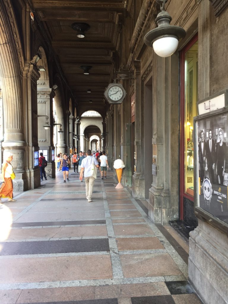 People walking beneath Bologna’s historic porticoes, with arched ceilings, stone columns, shopfronts, and a gelato cone sign along the covered walkway.