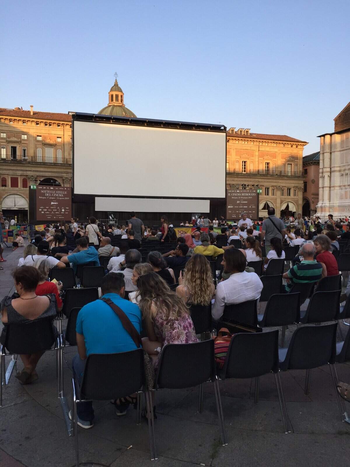 This lively outdoor movie event features audience watching film on big screen in historic city square during evening sunset, capturing cheerful atmosphere and architectural surroundings.