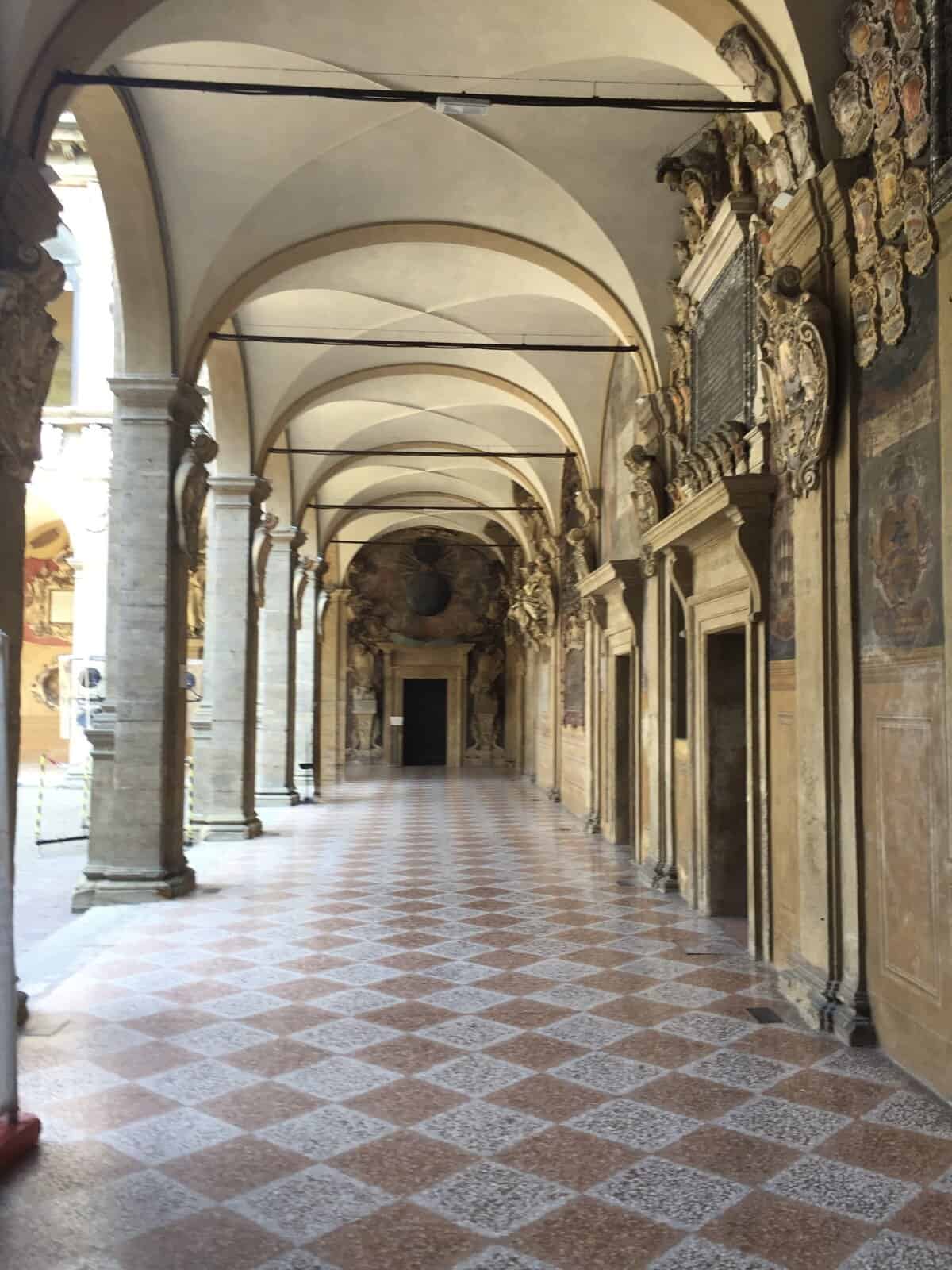 Covered portico walkway in Bologna, Italy, with arched ceilings, stone columns, and decorative frescoed walls stretching into the distance.
