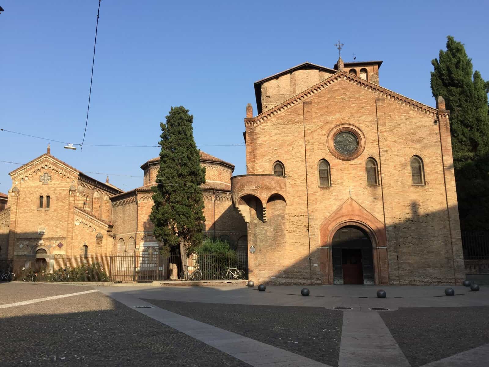 The Santo Stefano church complex in Bologna, Italy, showing warm brick Romanesque buildings and a quiet courtyard under a clear blue sky.