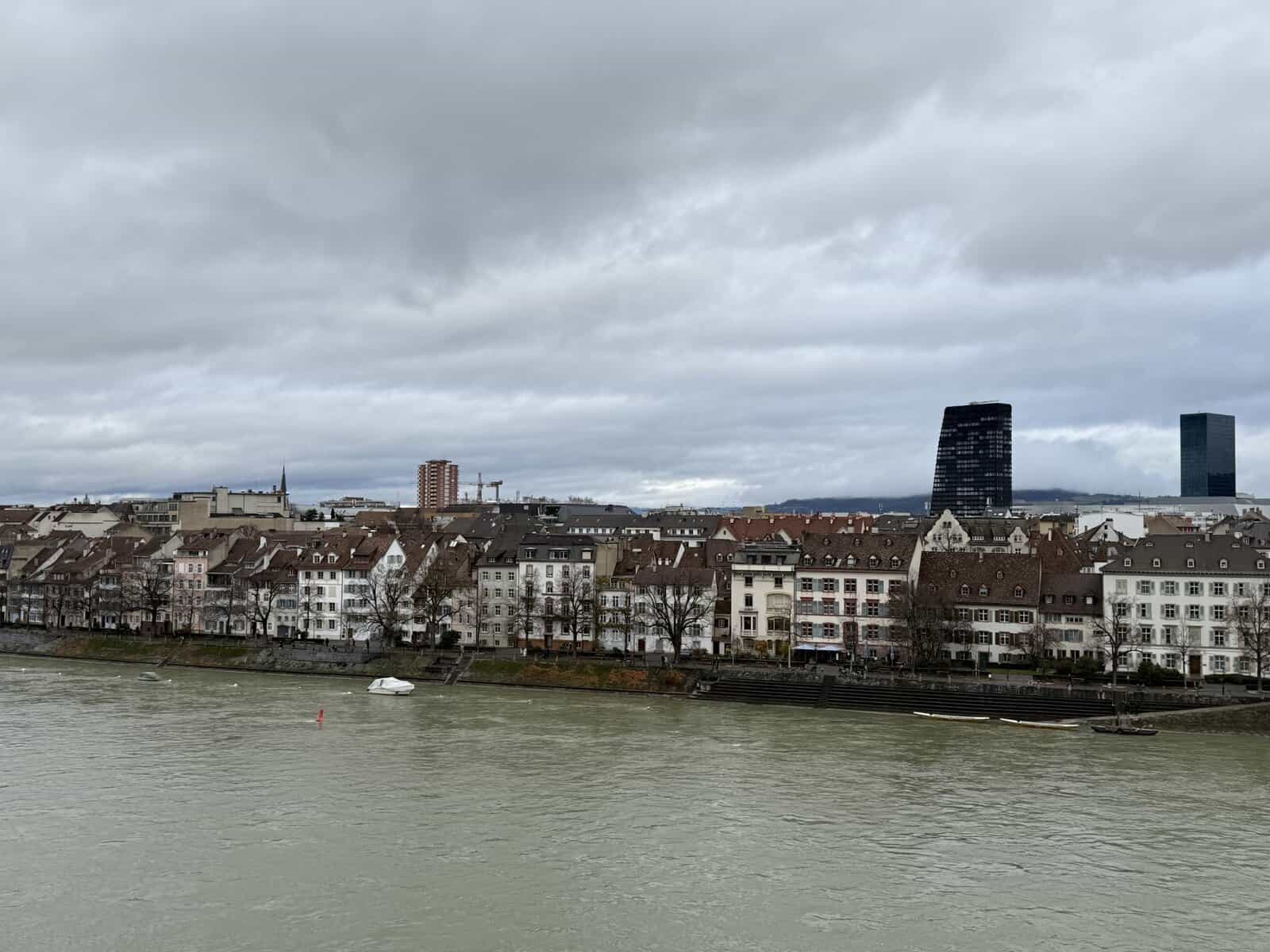 View across the Rhine River toward Kleinbasel in Basel Switzerland with historic riverside buildings and modern Roche towers under cloudy winter skies.