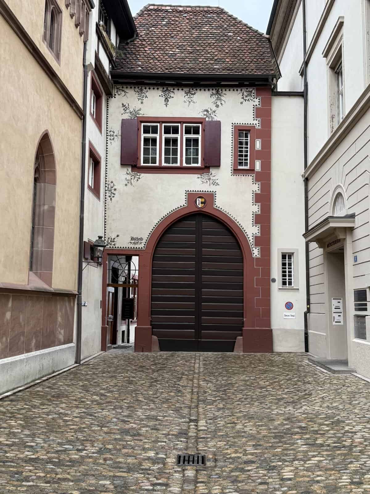 Historic decorated building in Basel Old Town featuring a painted façade, red sandstone trim, and large arched wooden doors along a cobblestone street.