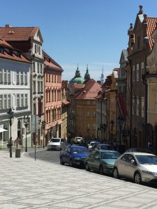 Historic street in Malá Strana Prague with colorful buildings and church domes in the distance.