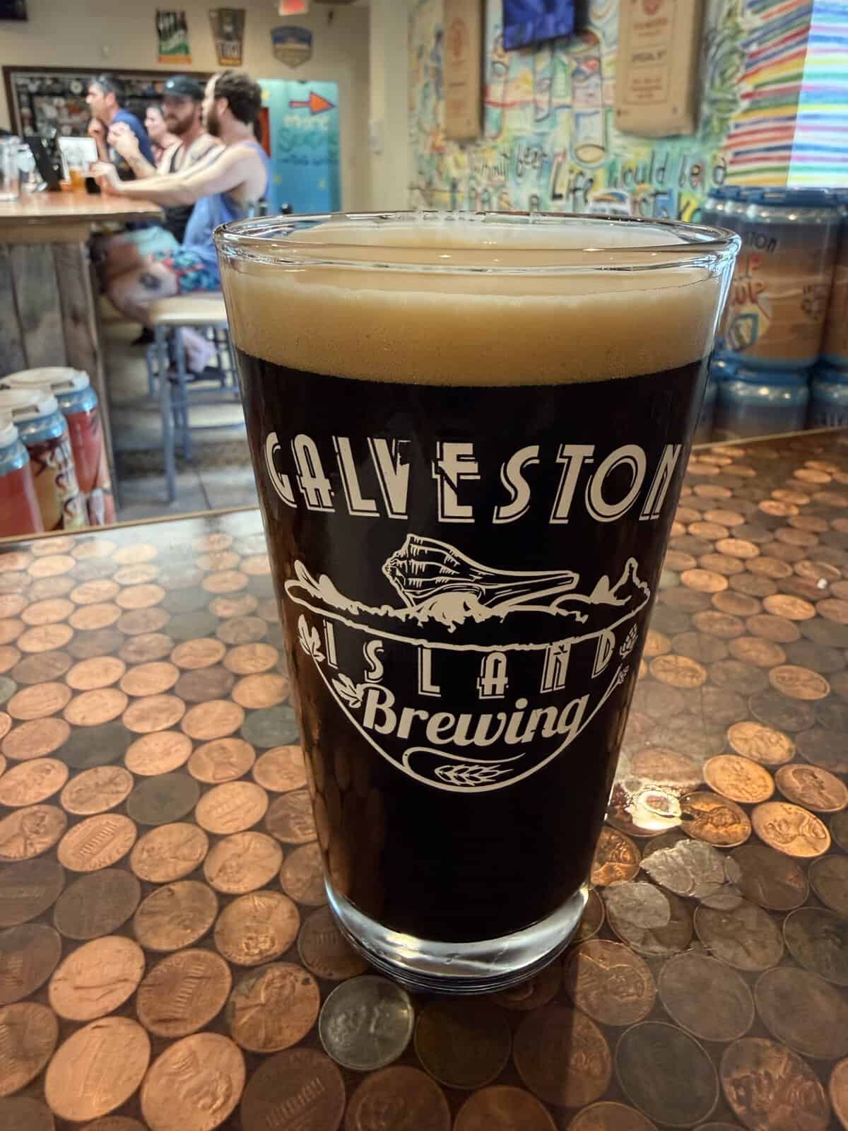 Dark beer at Galveston Island Brewing in Galveston, Texas with brewery logo on the glass and colorful brewery interior in the background.