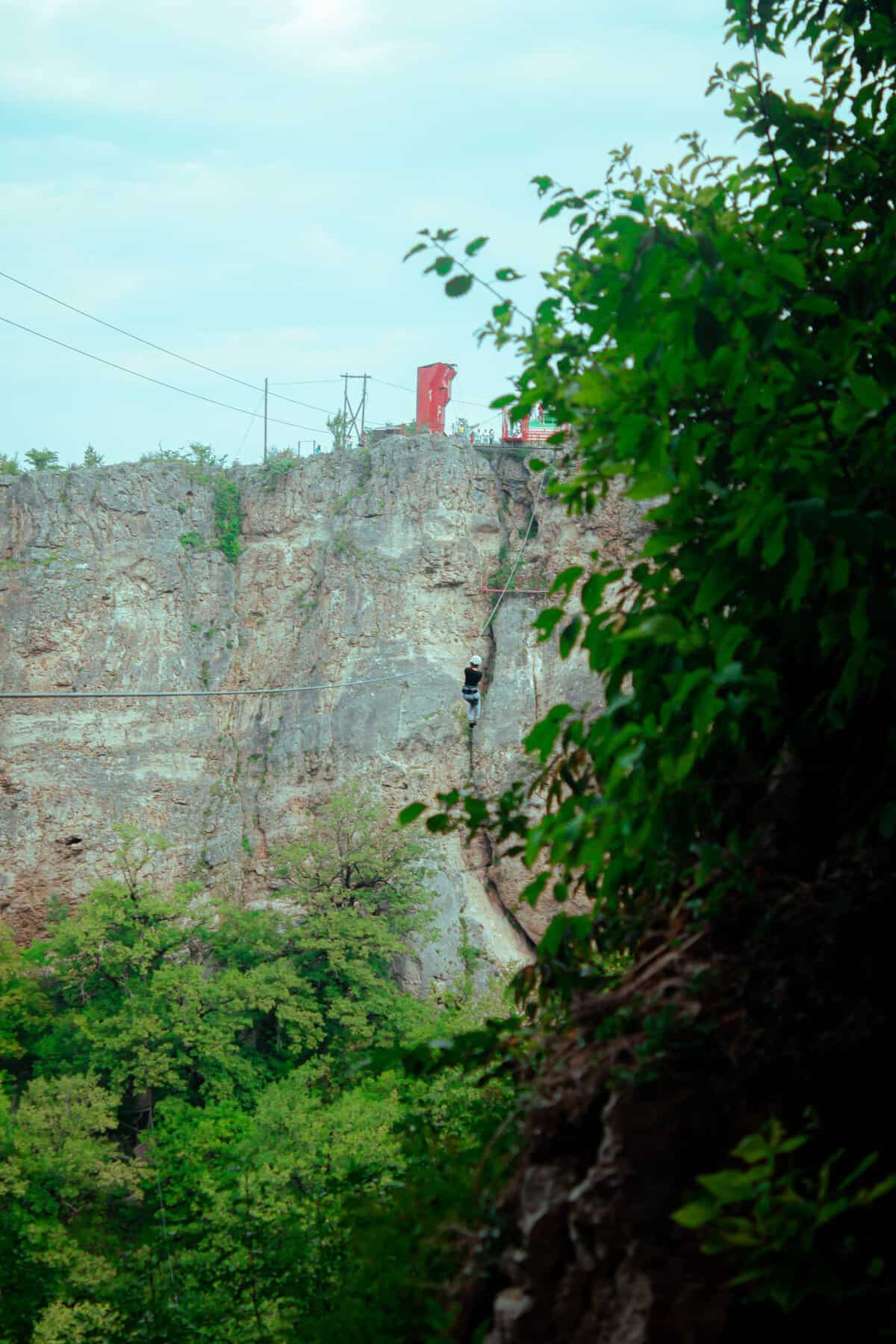 A thrilling view of an adventurer ziplining over a lush green canyon with rocky cliffs.