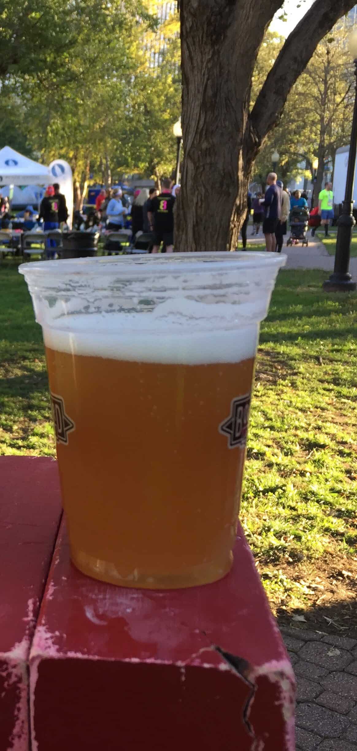Post race beer at the Kansas City Marathon finish festival with runners and race activities in the background.